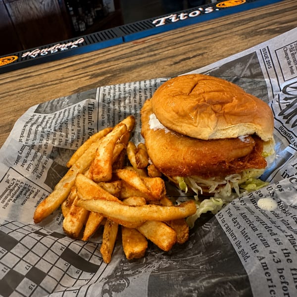 A fried fish sandwich with lettuce and tartar sauce, served with seasoned fries on newspaper print paper.