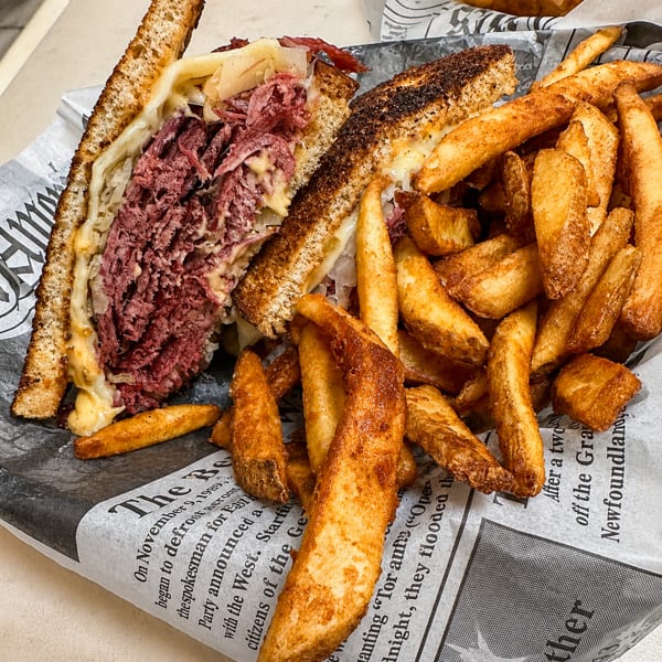 Toasted sandwich with corned beef, sauerkraut, and cheese, served with golden wedge fries on newspaper-print paper.