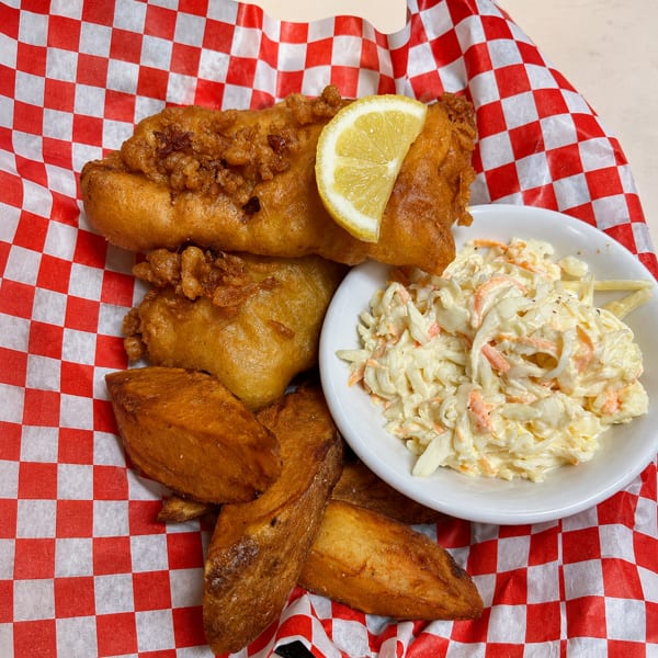 Two pieces of fried fish with a lemon, potato wedges, and coleslaw on checkered paper.