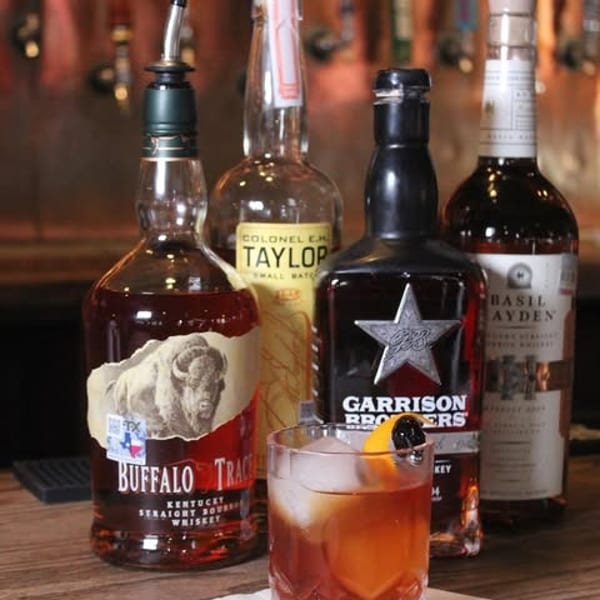 Four bourbon bottles and a garnished Old Fashioned cocktail on a wooden bar counter.